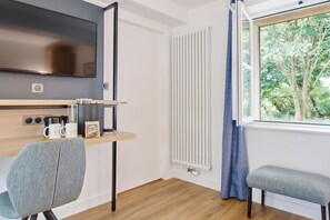 A modern room with a flat-screen TV, a wooden desk with a coffee maker, two mugs, and a bench by a window with a view of trees. - Les Maisons De Tatihou, The Originals Relais (Saint-Vaast-la-Hougue)