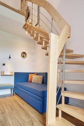 A modern living room with a blue sofa, a wooden staircase, and a wall-mounted mirror. - Les Maisons De Tatihou, The Originals Relais (Saint-Vaast-la-Hougue)