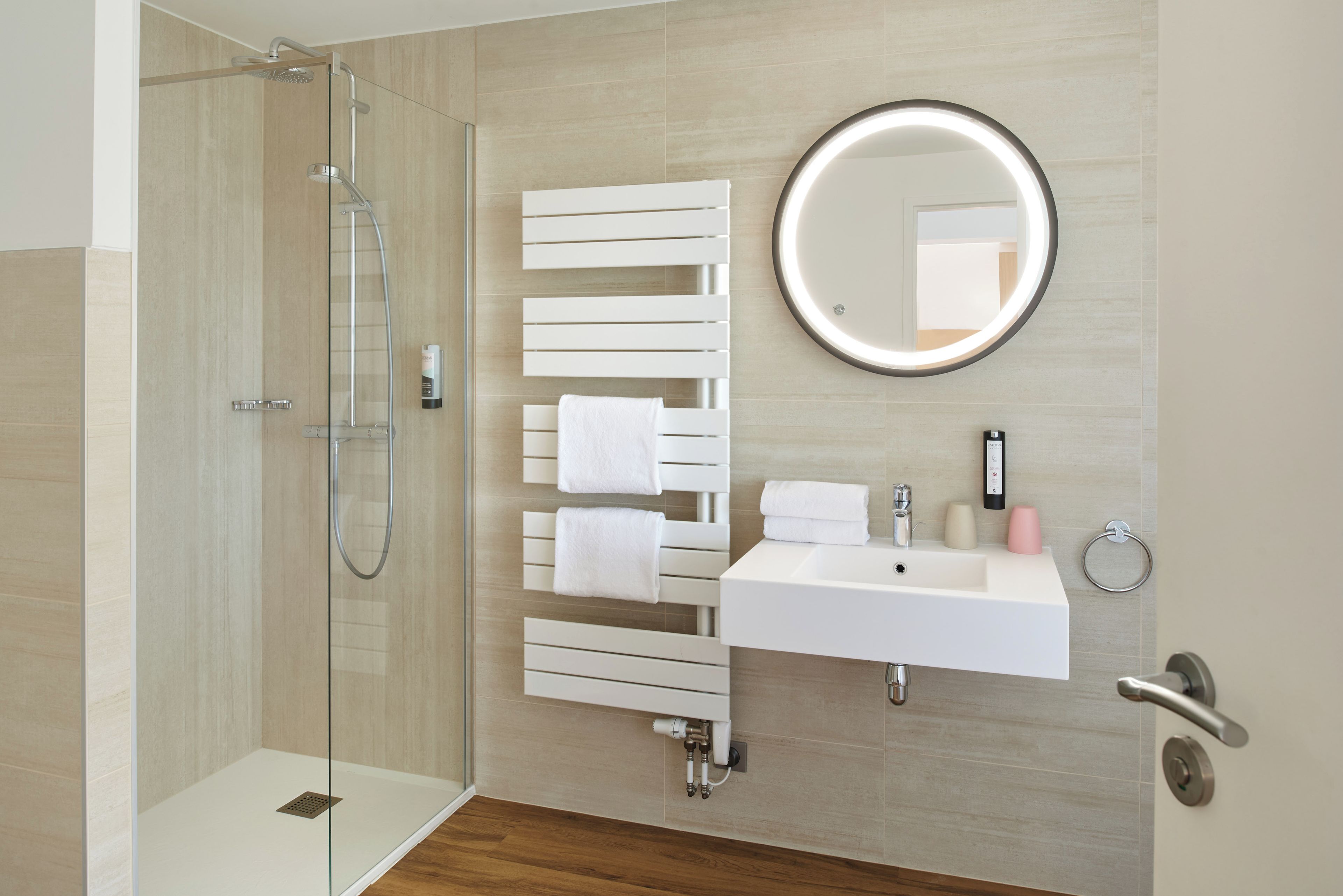 A modern bathroom with a glass-enclosed shower, a white sink, a round mirror, and a towel rack with multiple towels.