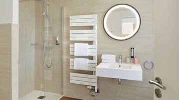 A modern bathroom with a glass-enclosed shower, a white sink, a round mirror, and a towel rack with multiple towels.