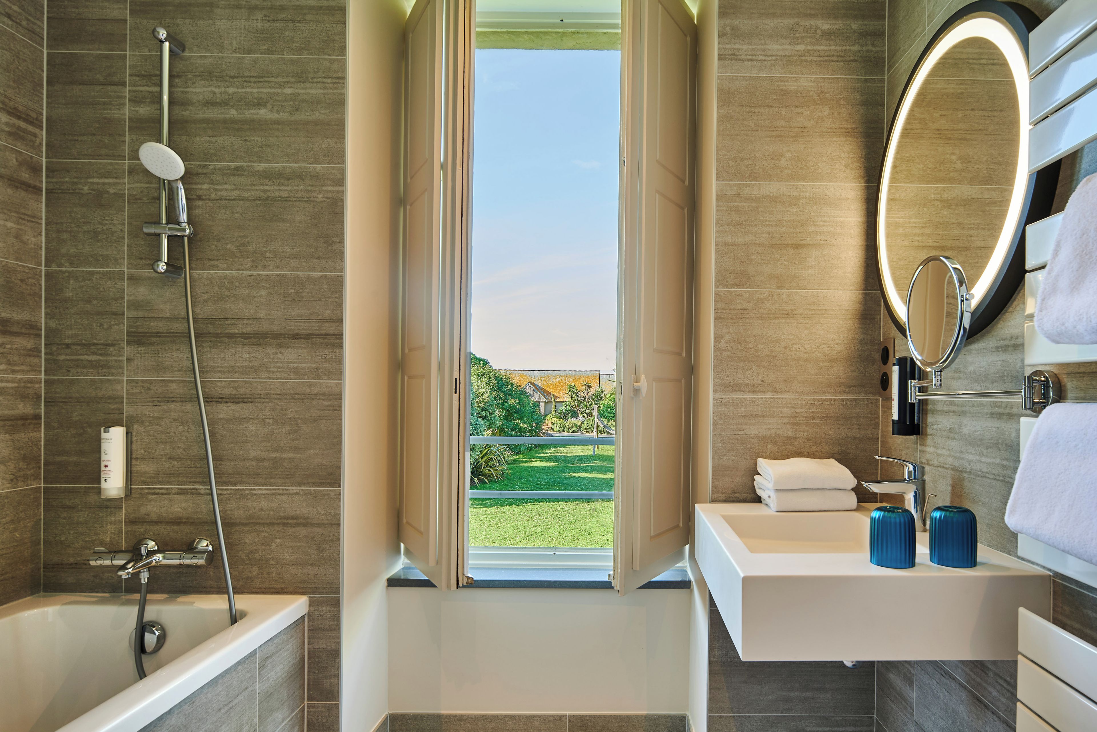 Een moderne badkamer met een badkuip, douche, wastafel met spiegel, en een raam met uitzicht op een groen landschap.