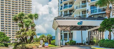 A modern hotel entrance with a covered walkway, palm trees, and a view of high-rise buildings in the background.