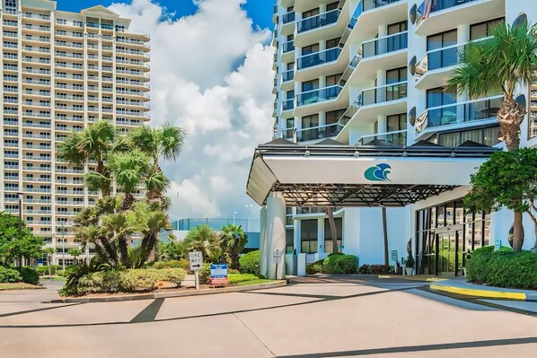 A modern hotel entrance with a covered walkway, palm trees, and a view of high-rise buildings in the background.