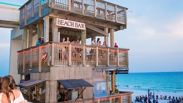 En träkonstruktion i flera våningar med skylten ”BEACH BAR”, ett räcke och utsikt över havet.