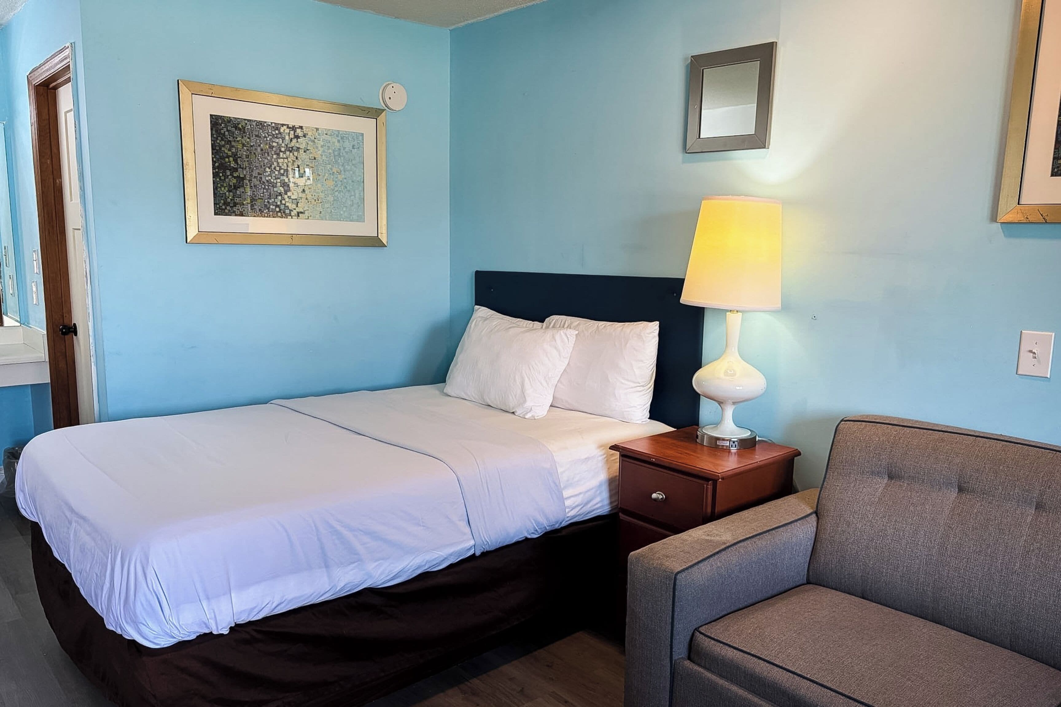 A hotel room with a bed, a nightstand with a lamp, a chair, and a wall-mounted picture.