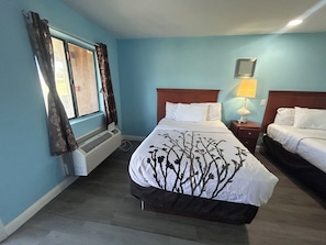 A hotel room with a bed, a bedside table with a lamp, a window with curtains, and a wall-mounted air conditioner.