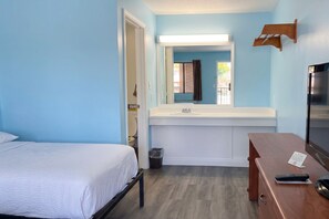 A hotel room with a bed, a desk, a mirror, a sink, and a trash bin.