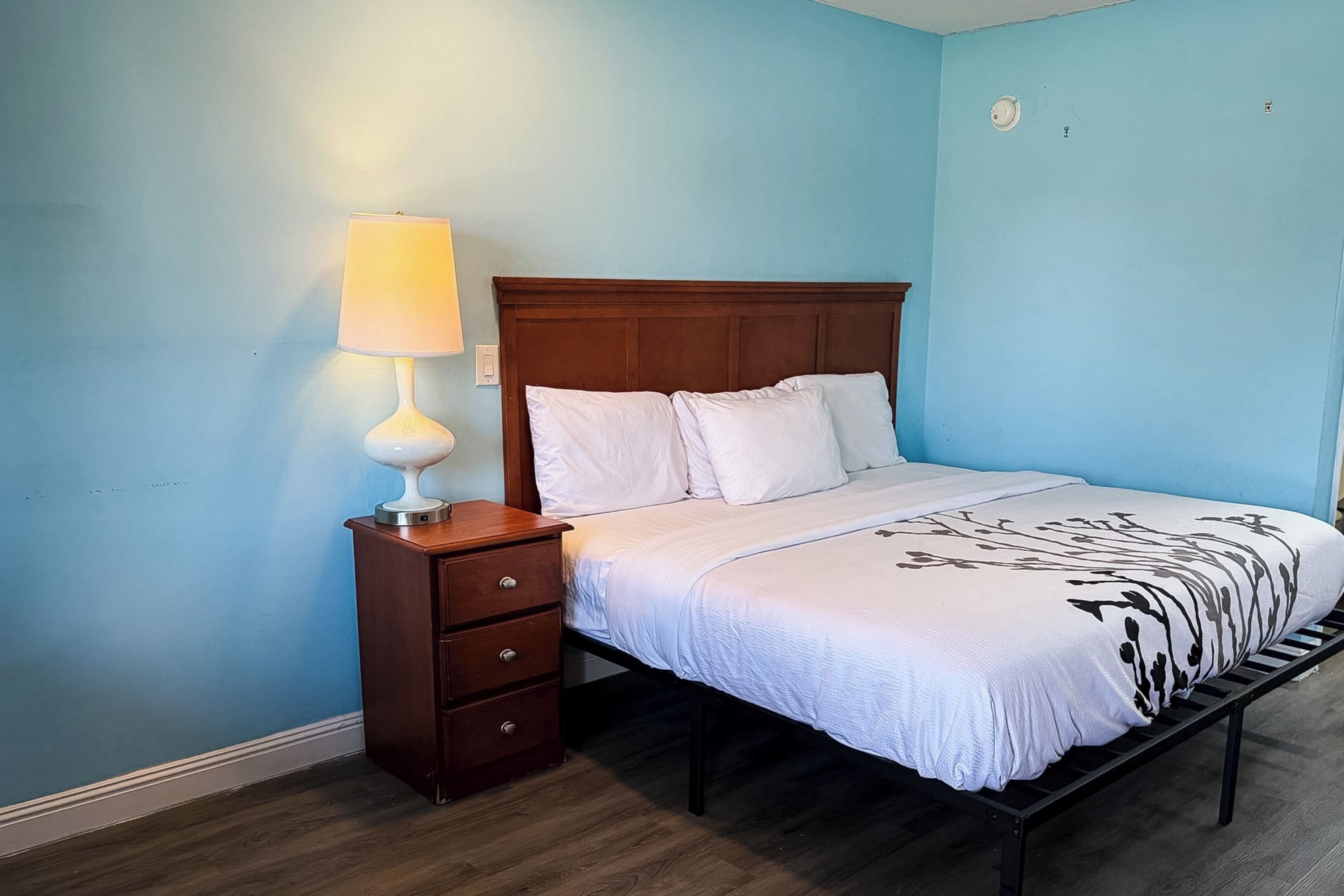 A bedroom with a wooden bed frame, white bedding, a bedside table with a lamp, and a blue wall.