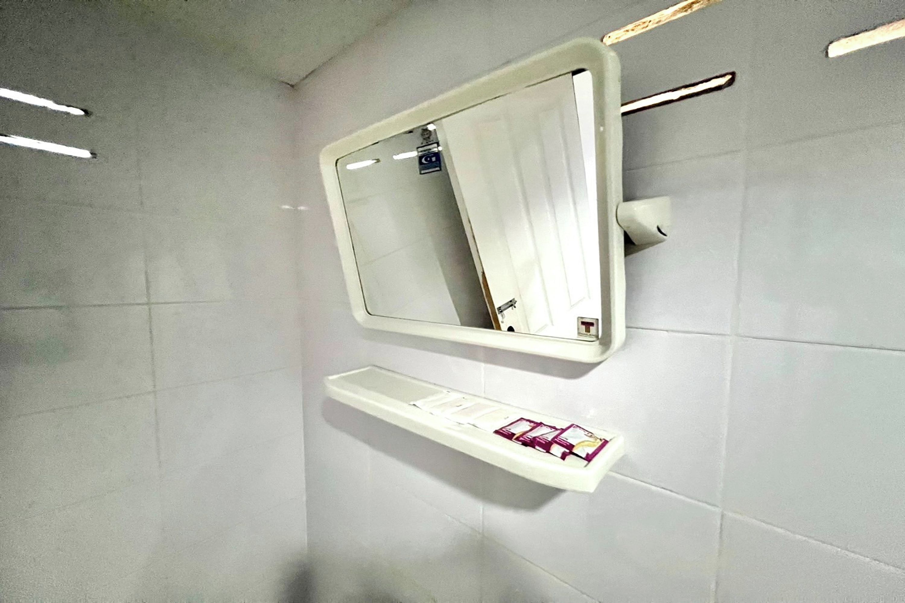 Casa de banho com parede revestida a azulejos brancos, um espelho e um prateleira com produtos de higiene pessoal.
