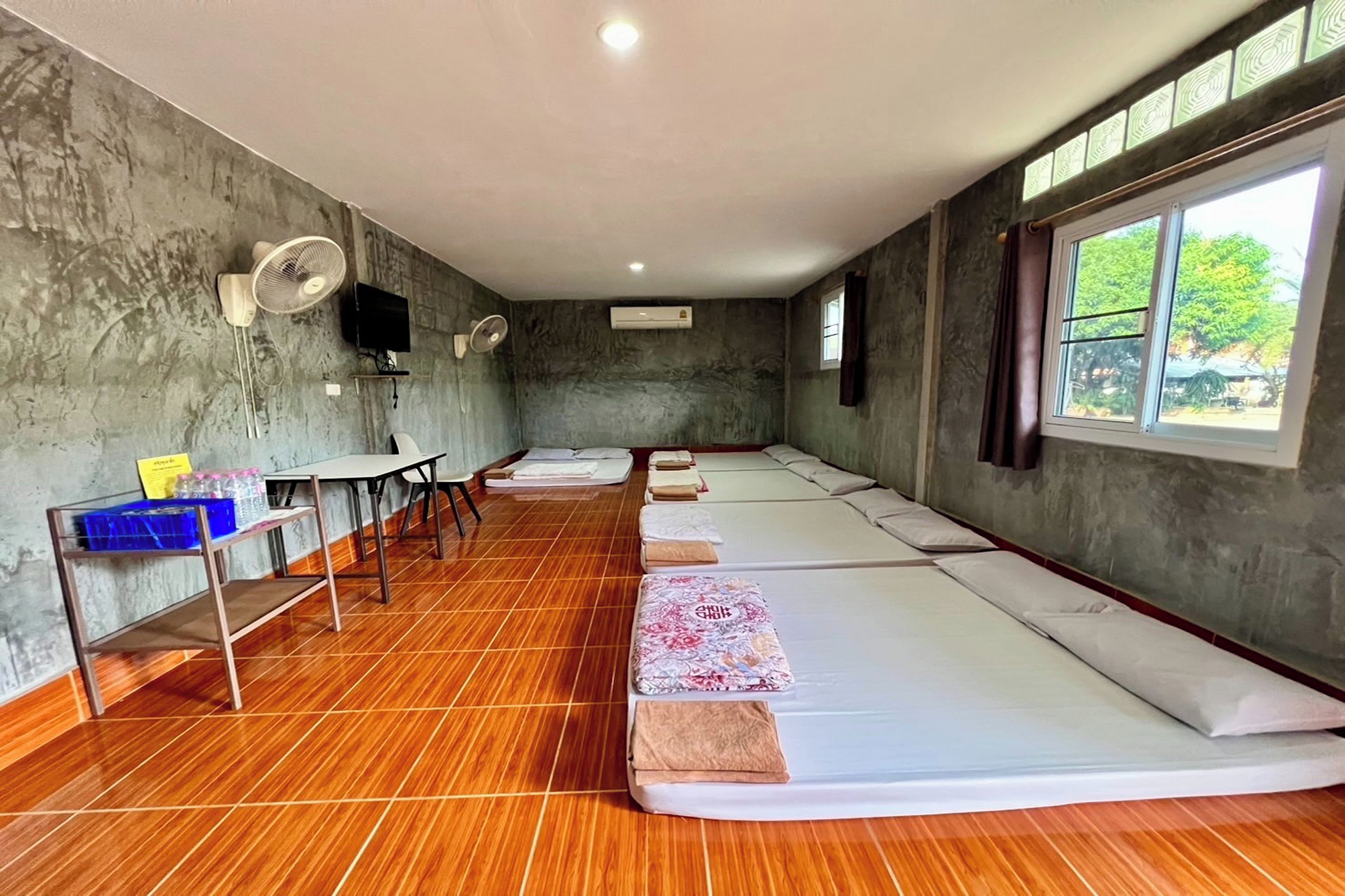 A room with multiple beds aligned along the wall, a table with a fan, and a window with a view of trees.