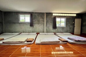 A room with four beds arranged in a row, each with a pillow and a folded towel. There are two windows with curtains, and a door is visible on the right. - Chalalai Resort Kham Chanot By Zuzu (Ban Dung)