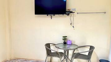A small room with a TV mounted on the wall, a small table with two chairs, a potted plant, and a trash bin.