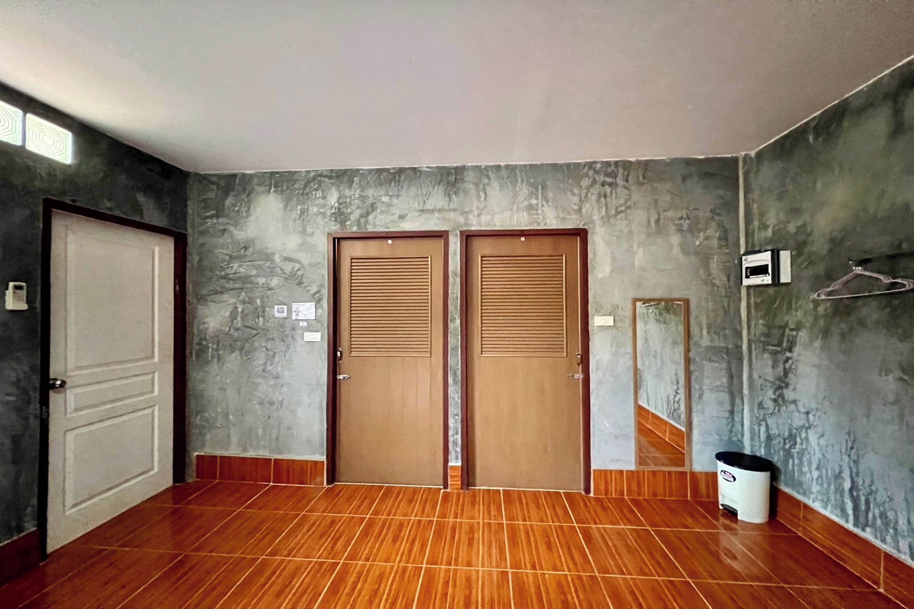 A room with two wooden doors, a tiled floor, a wall-mounted mirror, and a trash bin.