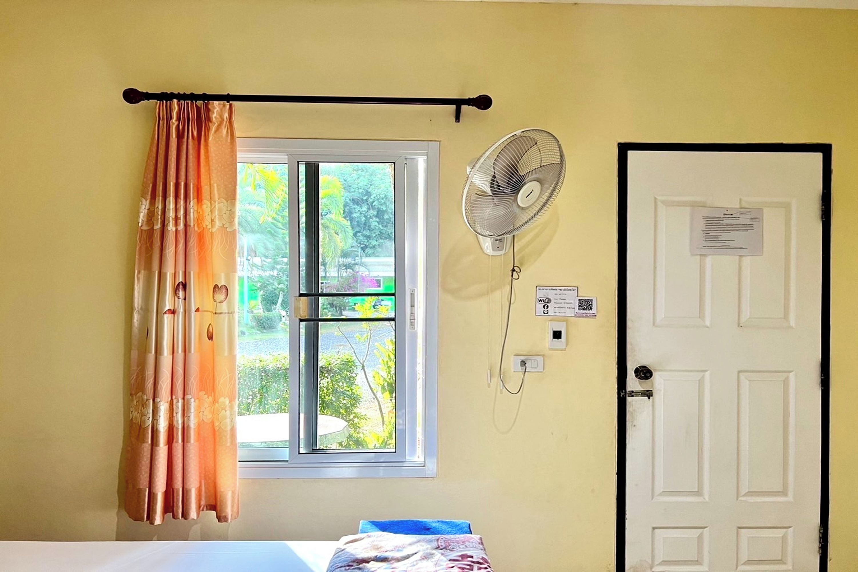 A room with a bed, a window with curtains, a wall-mounted fan, and a door with a notice.