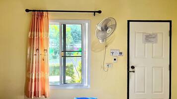A room with a bed, a window with curtains, a wall-mounted fan, and a door with a notice.