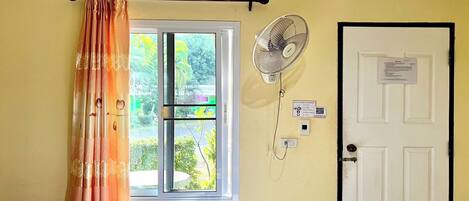 A room with a bed, a window with curtains, a wall-mounted fan, and a door with a notice.