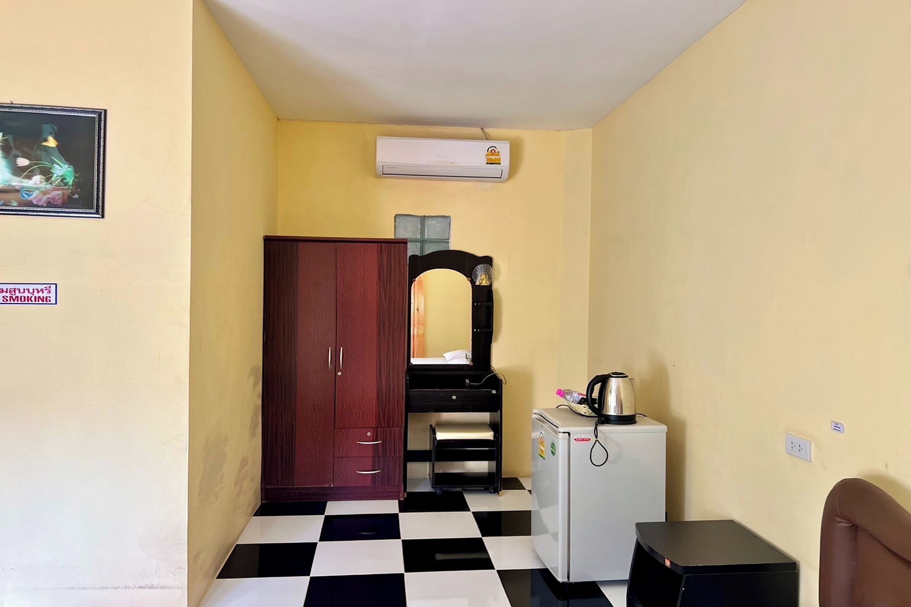 A small room with a checkered floor, a wooden wardrobe, a white refrigerator, and a small table with a kettle.
