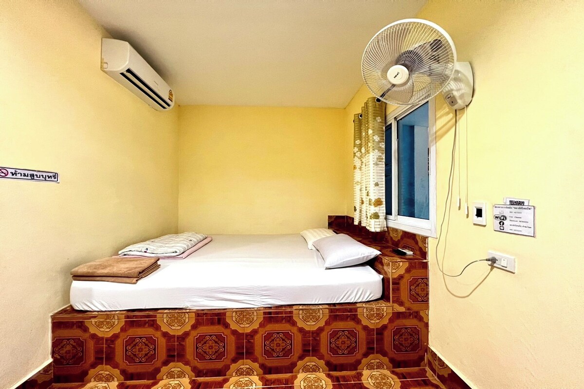 A small, single-bed room with a patterned floor, a standing fan, an air conditioning unit, a window with curtains, and a sign on the wall.
