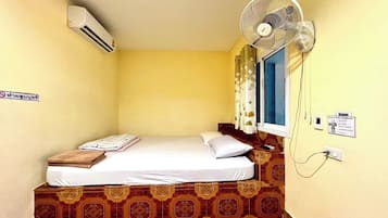 Une petite chambre avec un lit simple, un sol à motifs, un ventilateur sur pied, un climatiseur, une fenêtre avec des rideaux et un panneau affiché sur le mur.