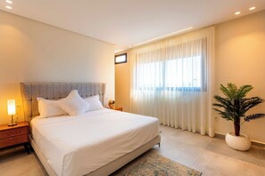 Ein modernes Schlafzimmer mit einem großen Bett, einem Nachttisch mit Lampe, einer Zimmerpflanze und einem Fenster mit transparenten Vorhängen.