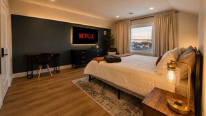 Et moderne soveværelse med en stor seng, et skrivebord, et fjernsyn der viser 'NETFLIX' og udsigt ud i det fri gennem et vindue.