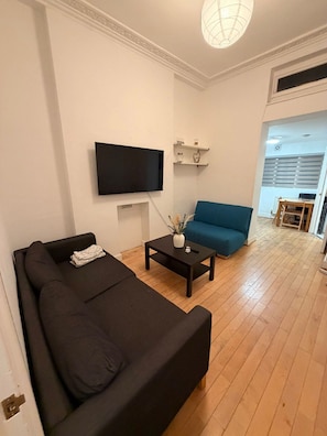 A living room with a black sofa, a blue sofa, a coffee table, a flat-screen TV mounted on the wall, and a small shelf with decorative items.