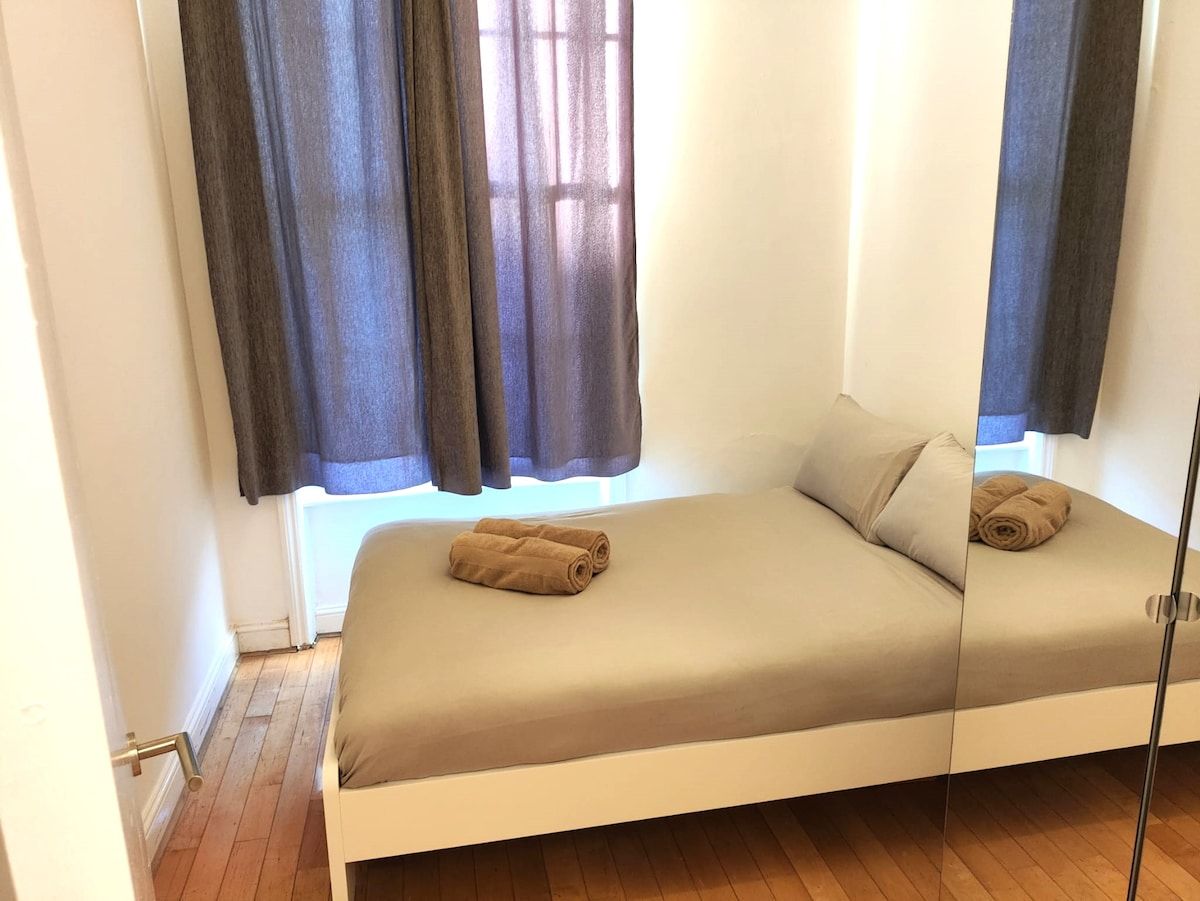 A single bed with a beige mattress and pillows, a window with curtains, and a wooden floor.