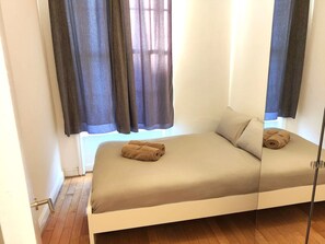 A single bed with a beige mattress and pillows, a window with curtains, and a wooden floor.