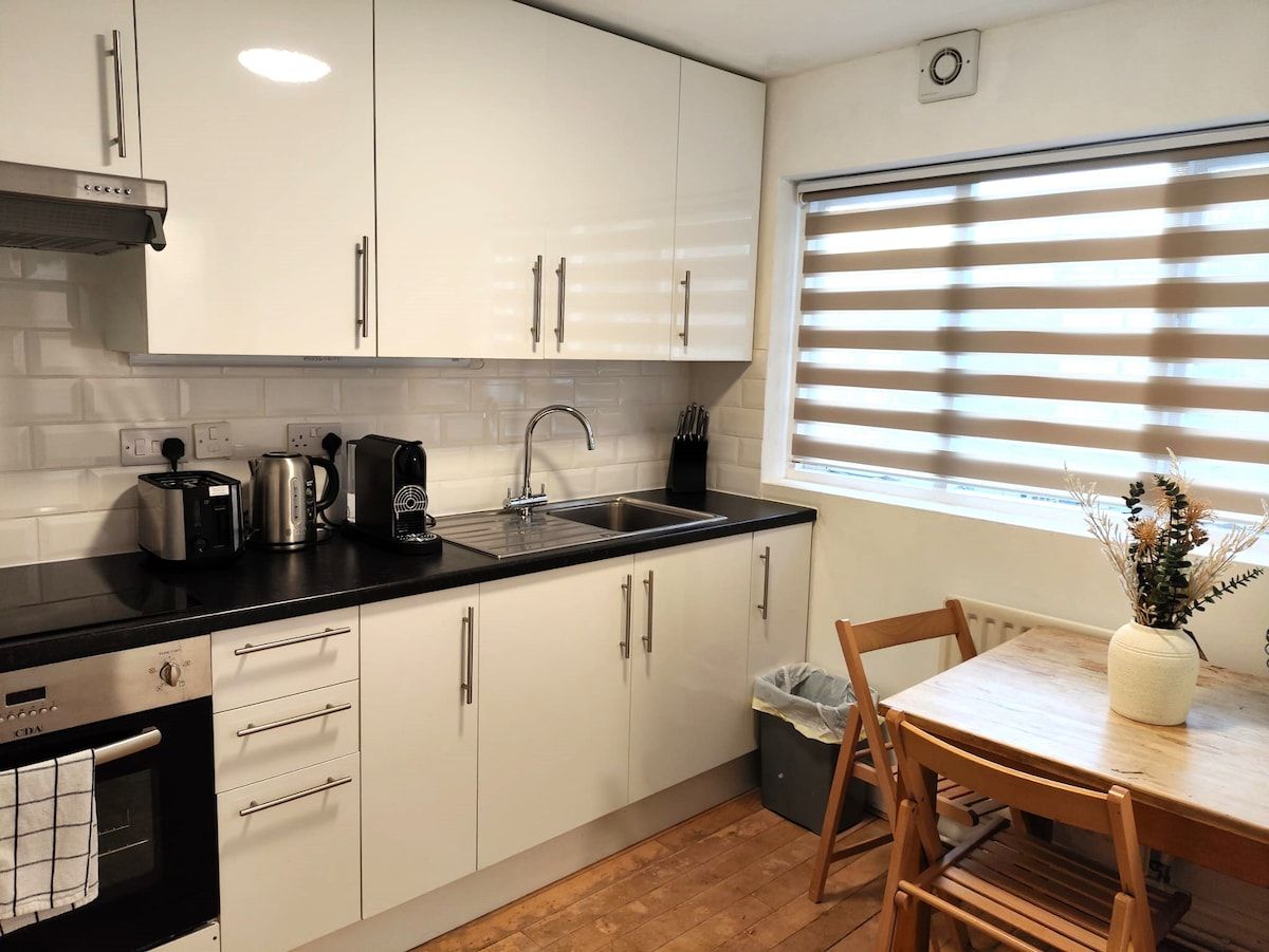 A compact kitchen with white cabinets, a black countertop, a sink, a toaster, a kettle, and a window with blinds.