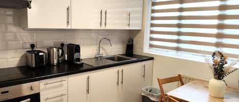 A compact kitchen with white cabinets, a black countertop, a sink, a toaster, a kettle, and a window with blinds.