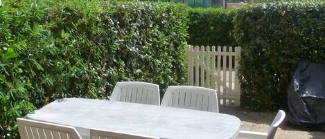 A patio area with a table and chairs, surrounded by greenery and a white picket fence.