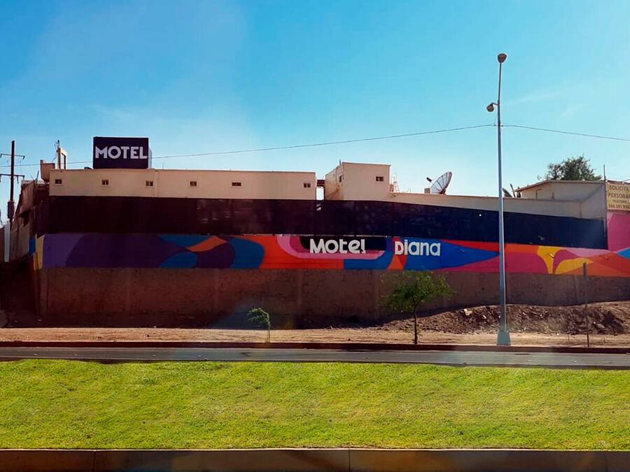 Un motel orné d’une fresque colorée sur le côté, un ciel d’un bleu éclatant et une zone herbeuse au premier plan.