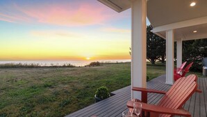 Une terrasse en bois avec des chaises, une vue sur le coucher de soleil sur l’océan et une zone herbeuse.
