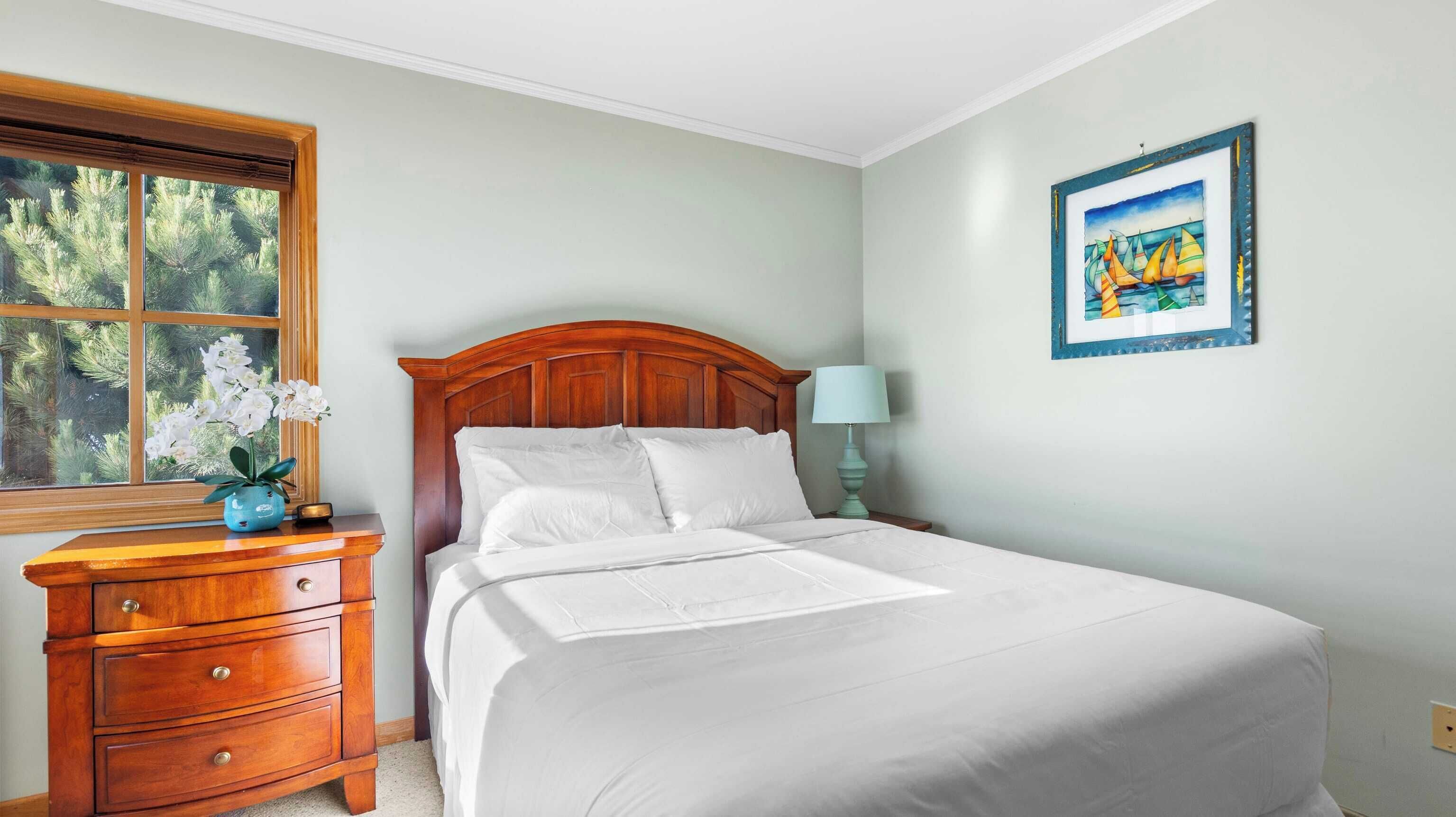 A neatly made bed with white linens, a wooden nightstand with a lamp, and a window with a view of trees.