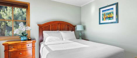 A neatly made bed with white linens, a wooden nightstand with a lamp, and a window with a view of trees.