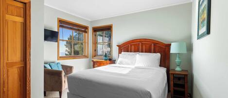 Une chambre à coucher avec un grand lit, deux fenêtres, une télévision, une chaise, une table de chevet et une lampe.