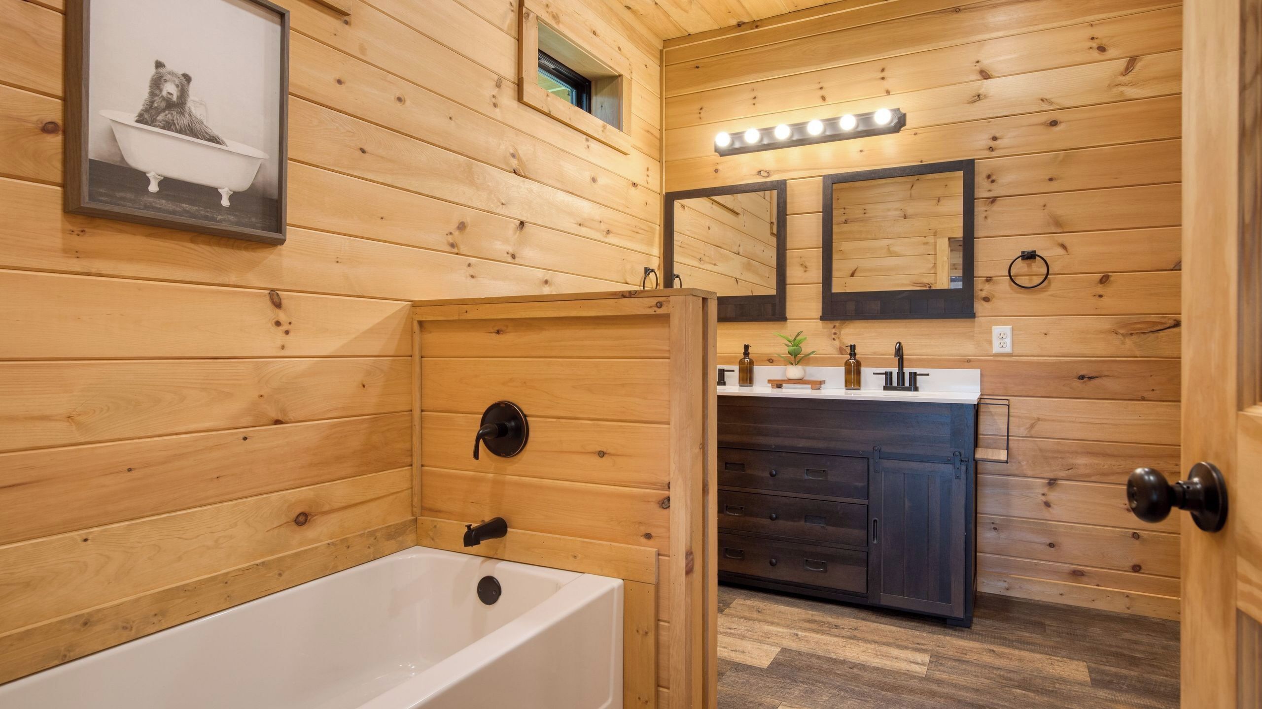 A wooden cabin bathroom with a bathtub, a sink with a mirror, and a framed photo on the wall.