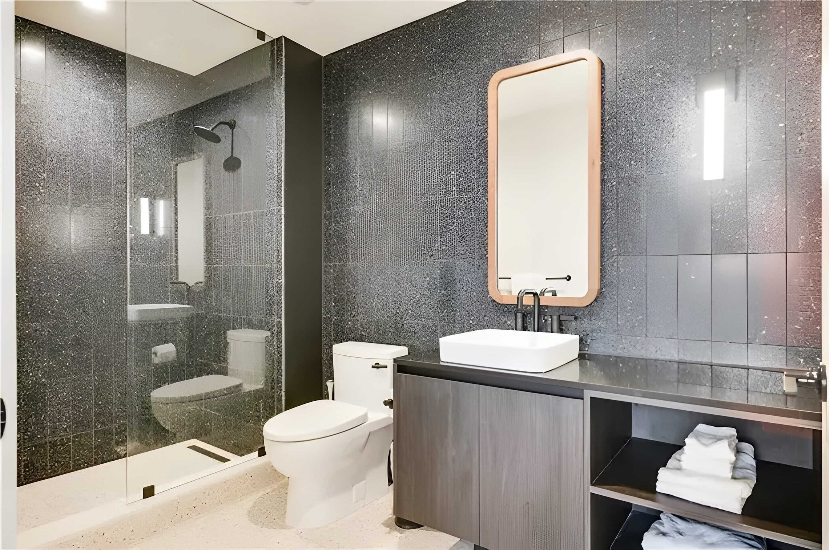 Et moderne badeværelse med en glasafskærmet brusekabine, et hvidt toilet, en vask med spejl og et træskab.