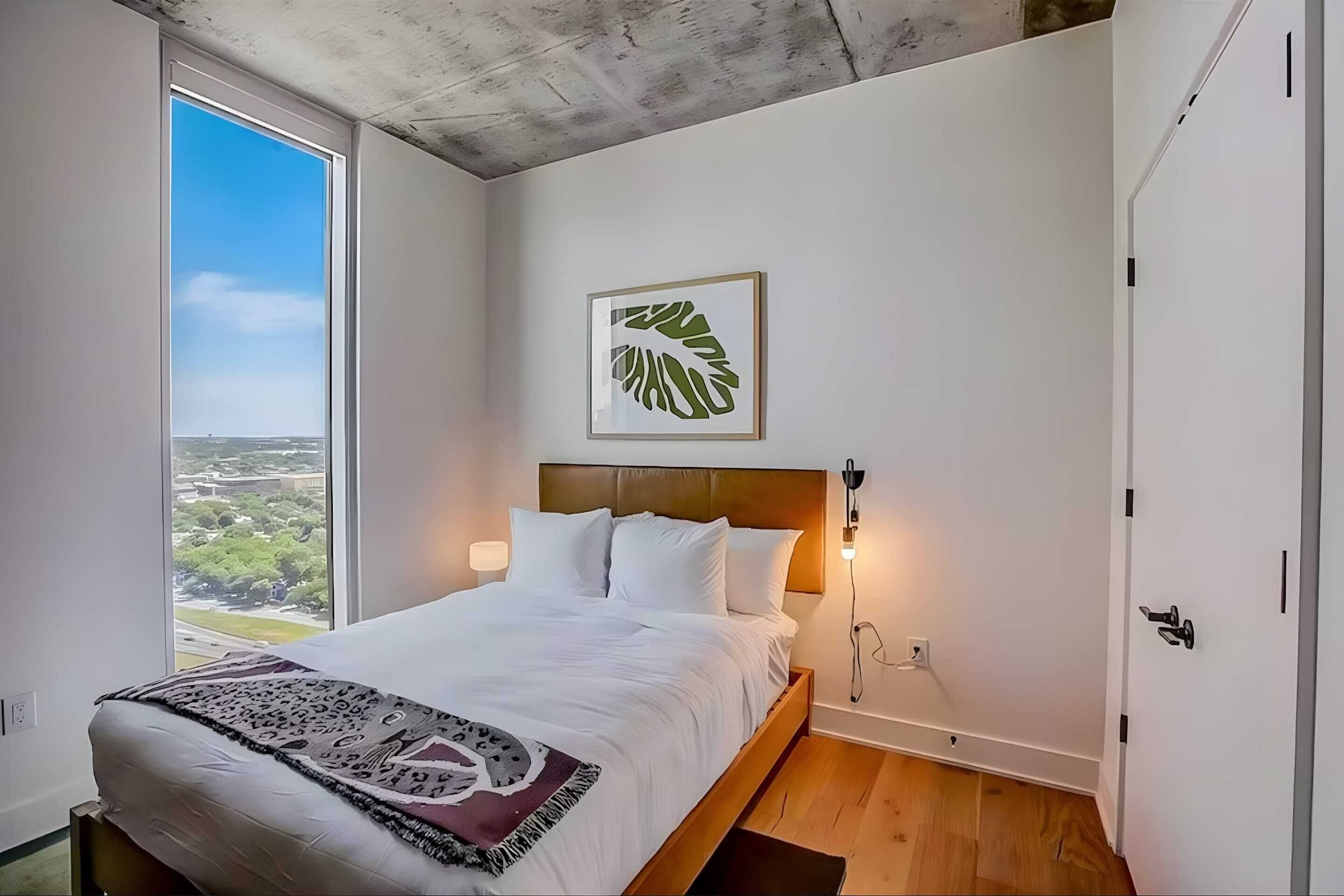 Un dormitorio con cama, vista al exterior, una obra de arte enmarcada y piso de madera.