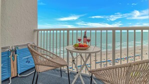 A balcony with wicker chairs, a table with fruit and wine glasses, and a view of the beach.