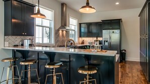 Cocina moderna con gabinetes oscuros, una isla grande con taburetes de bar, electrodomésticos de acero inoxidable e iluminación colgante.