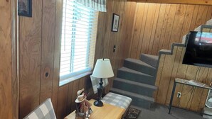 A cozy cabin interior with a wooden interior, a checkered bench, a small table with a lamp, a television, and a staircase.