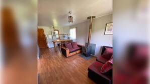 A spacious living area with a wooden floor, a sofa set with red cushions, a fireplace, and a kitchen in the background. - cabañas nuevo amanecer paillaco valdivia (Paillaco)
