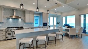Une cuisine moderne dotée d’un grand îlot central, de tabourets de bar, d’appareils électroménagers en acier inoxydable et de luminaires suspendus.