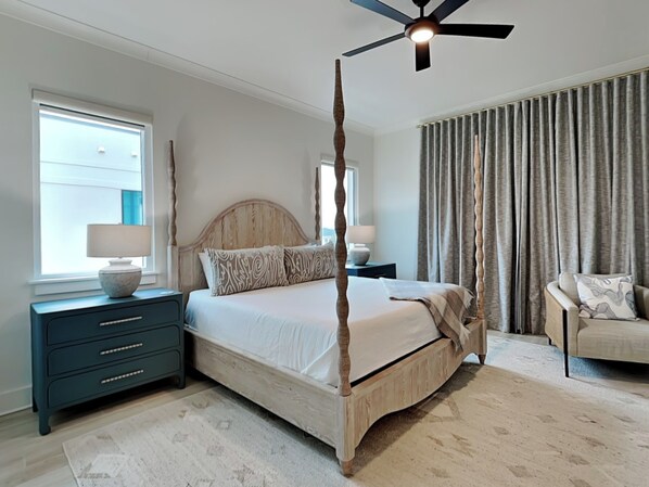 A bedroom with a four-poster bed, a ceiling fan, a nightstand with a lamp, a dresser, and a chair.