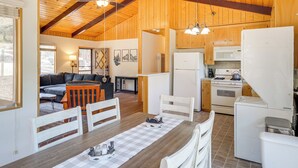 A cozy cabin-style kitchen with wooden beams, a dining area with a checkered tablecloth, and a living room with a sofa and fireplace. - Mountain-View Haven w/ Deck in Pine Mountain Club (Frazier Park)