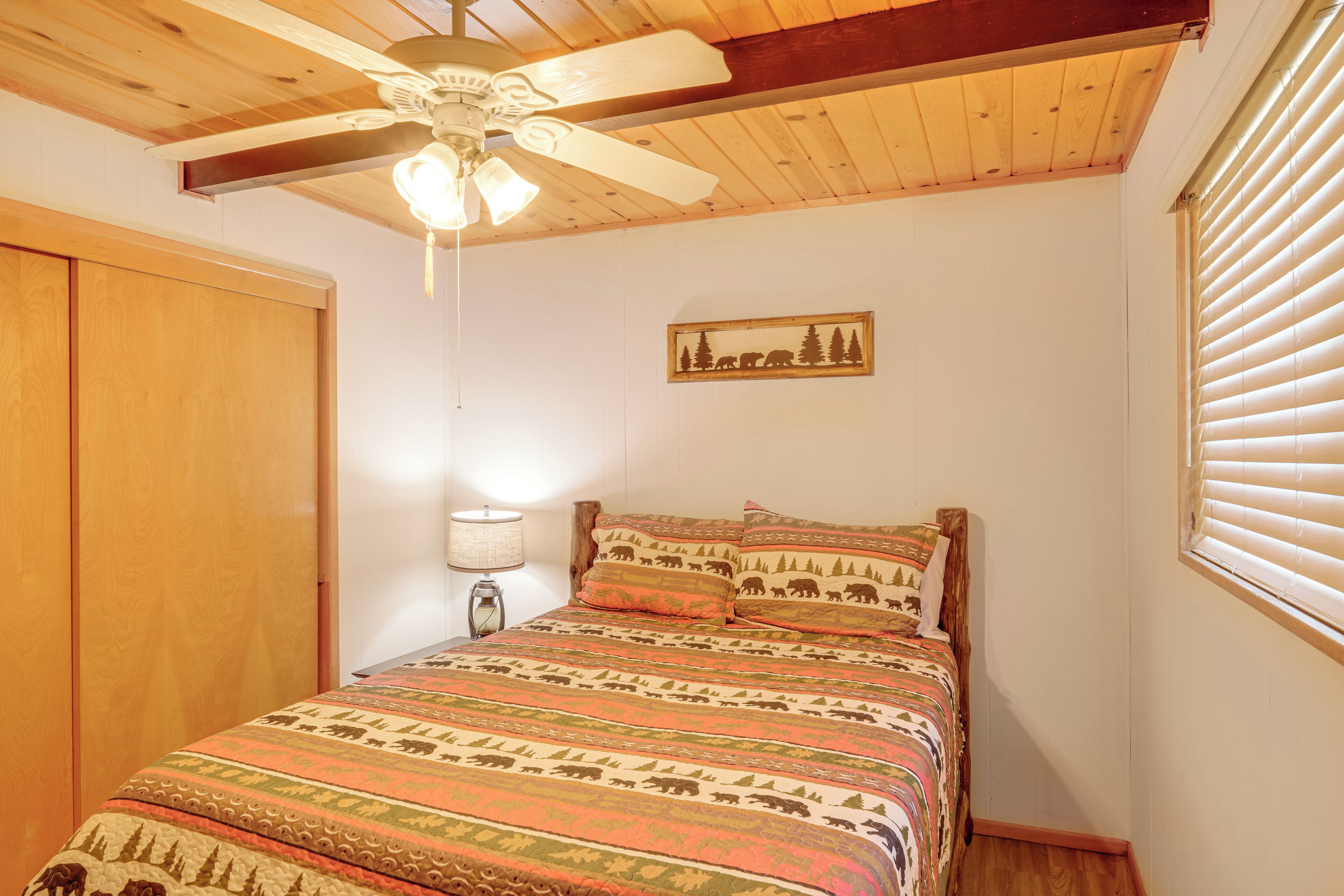 Une chambre à coucher comprenant un lit, un ventilateur de plafond, une table de chevet, une lampe, une fenêtre avec des stores et une porte.