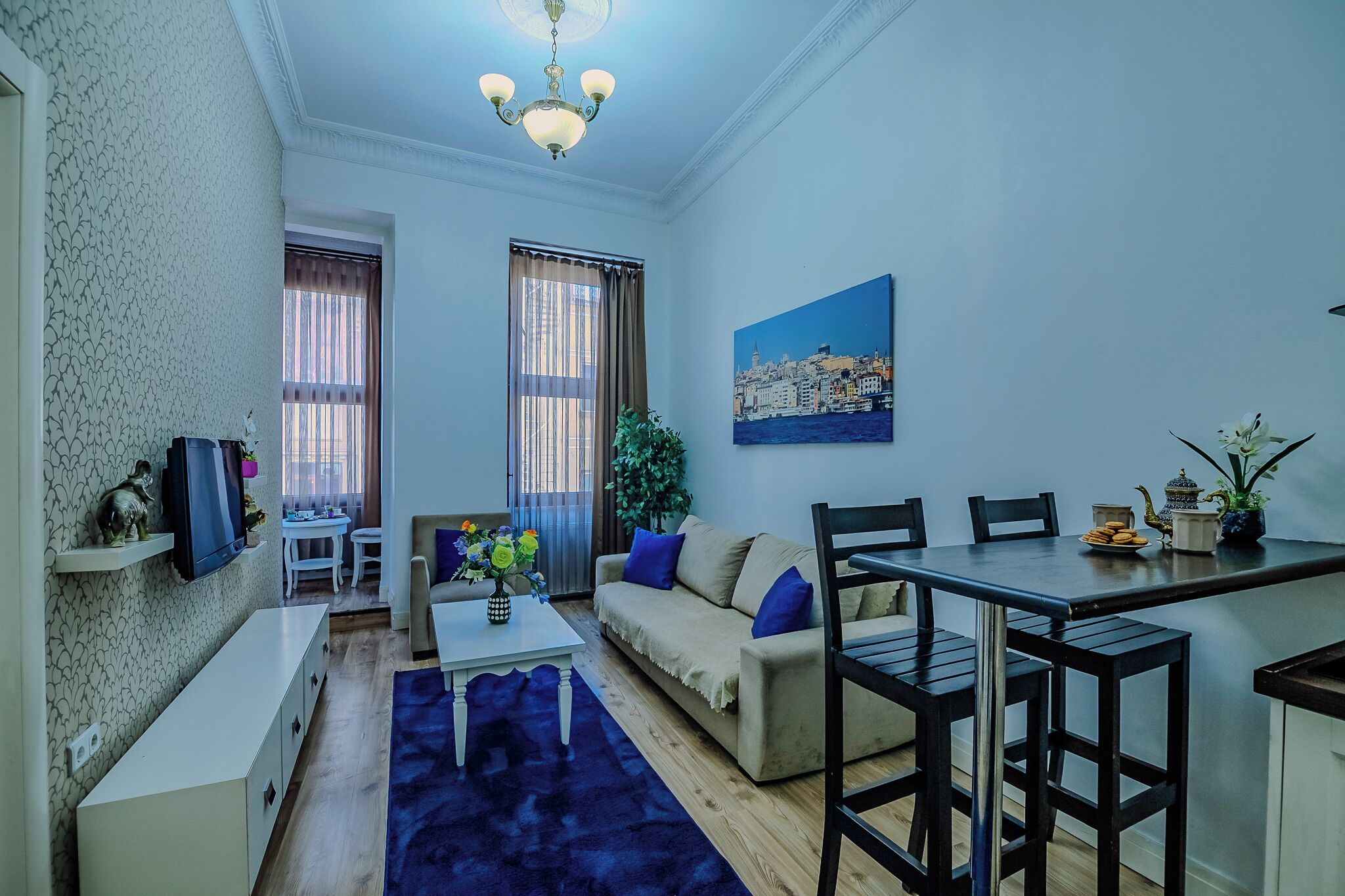 A living room with a sofa, coffee table, and dining area. There is a painting on the wall and a plant on the windowsill.