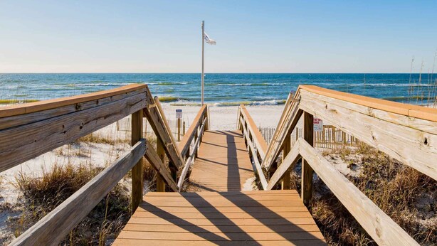 Une promenade en bois mène à une plage où se trouvent un mât et un panneau indiquant : « Restez sur la promenade ».