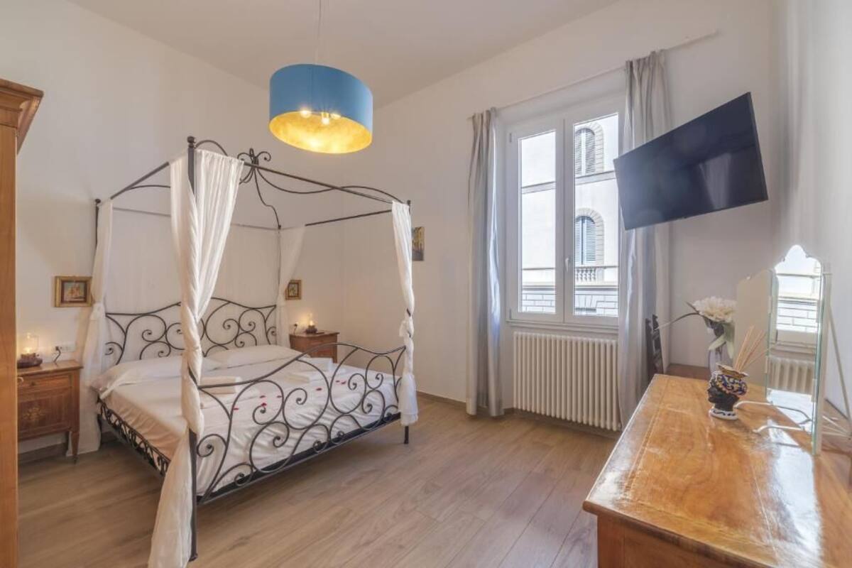 A bedroom with a four-poster bed, a wooden nightstand, a bedside table with a lamp, a large window with curtains, and a flat-screen TV mounted on the wall.